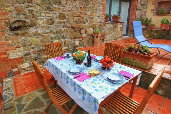 Un área de comedor al aire libre con paredes de piedra, una mesa dispuesta para dos personas y sillas de madera alrededor.