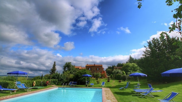 A swimming pool with blue lounge chairs and umbrellas, surrounded by greenery and a building in the background.