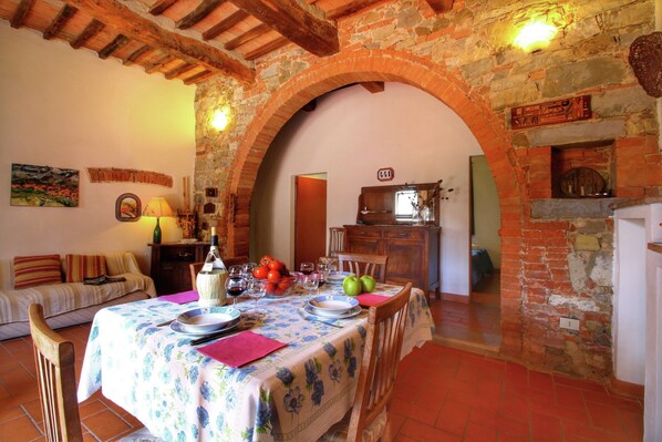 A dining area with a table set for two, a stone wall, and a rustic kitchen in the background.