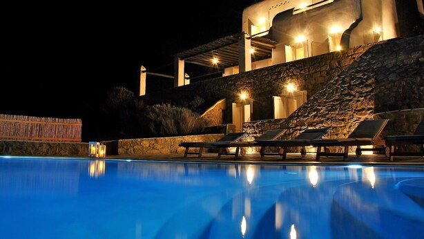 Vista noturna de um hotel com piscina, cadeiras de descanso e um muro de pedra.