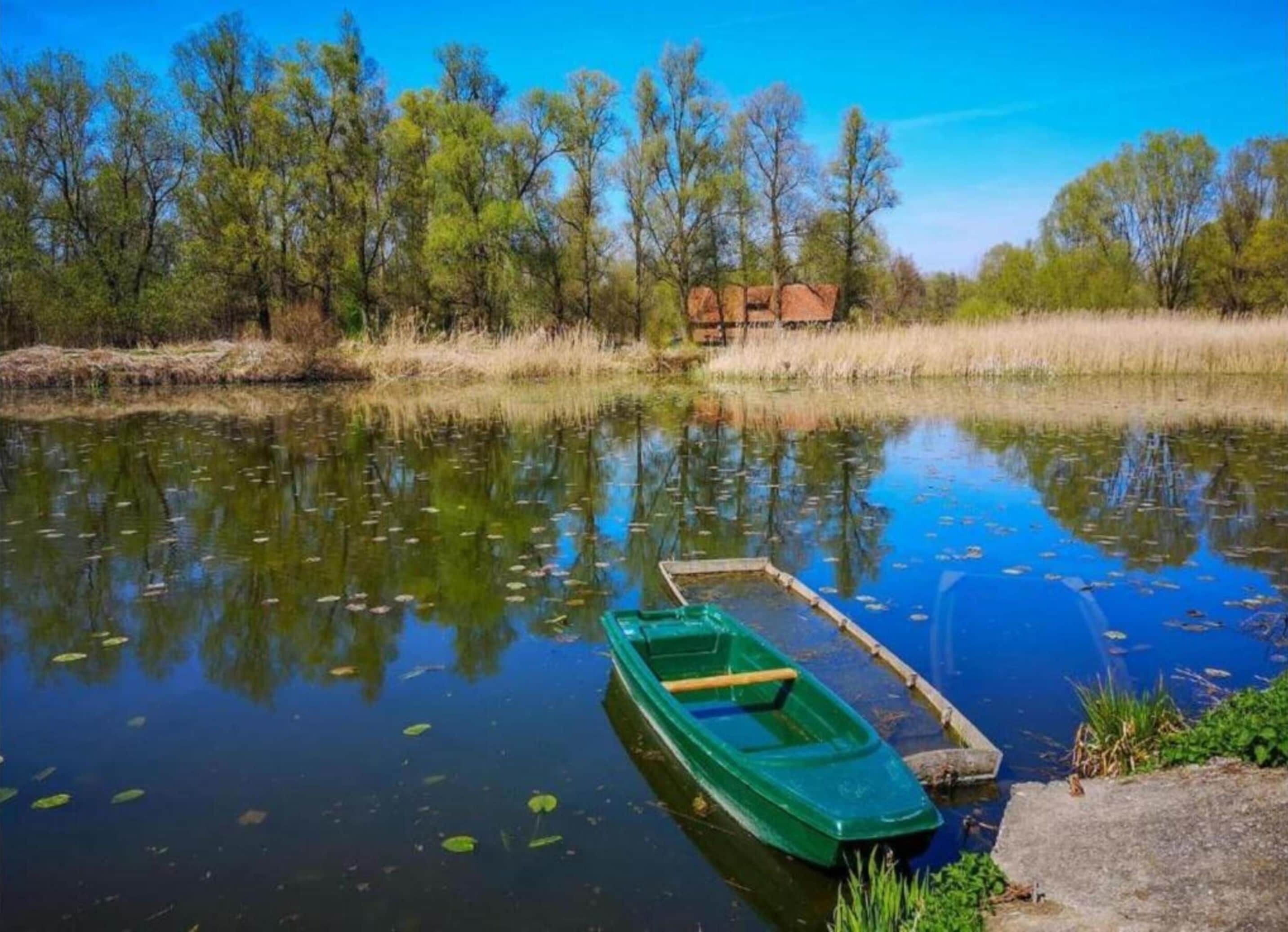 A calm river with a green rowboat docked on the right, surrounded by trees and a clear sky.