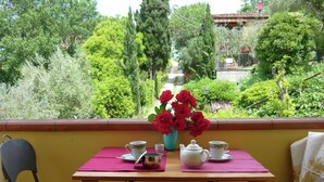Un balcone con un tavolo apparecchiato per due persone, un vaso di fiori rossi e vista su un giardino e un edificio sullo sfondo.
