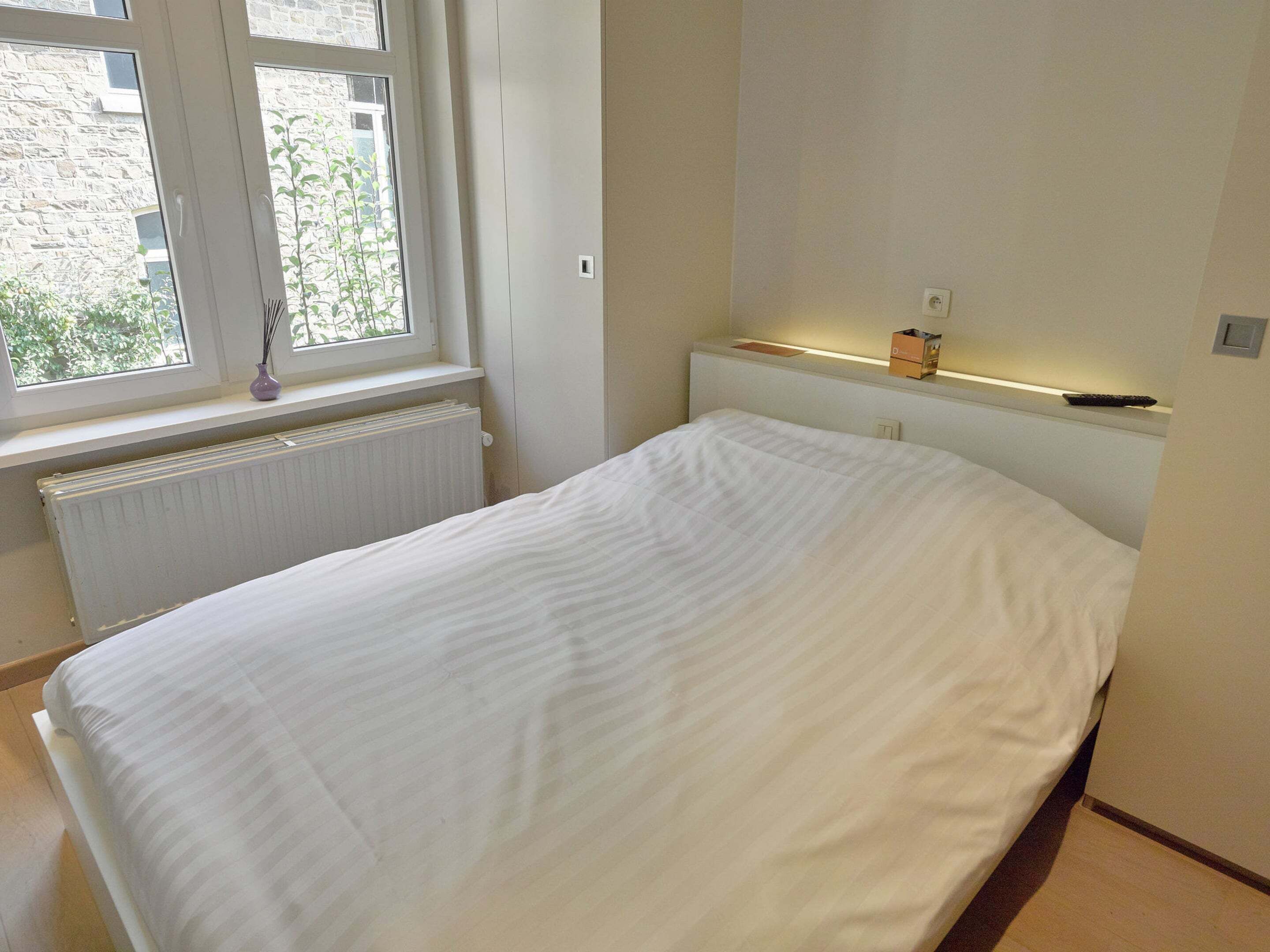 A bedroom with a bed, a window, a radiator, and a shelf with a remote control.