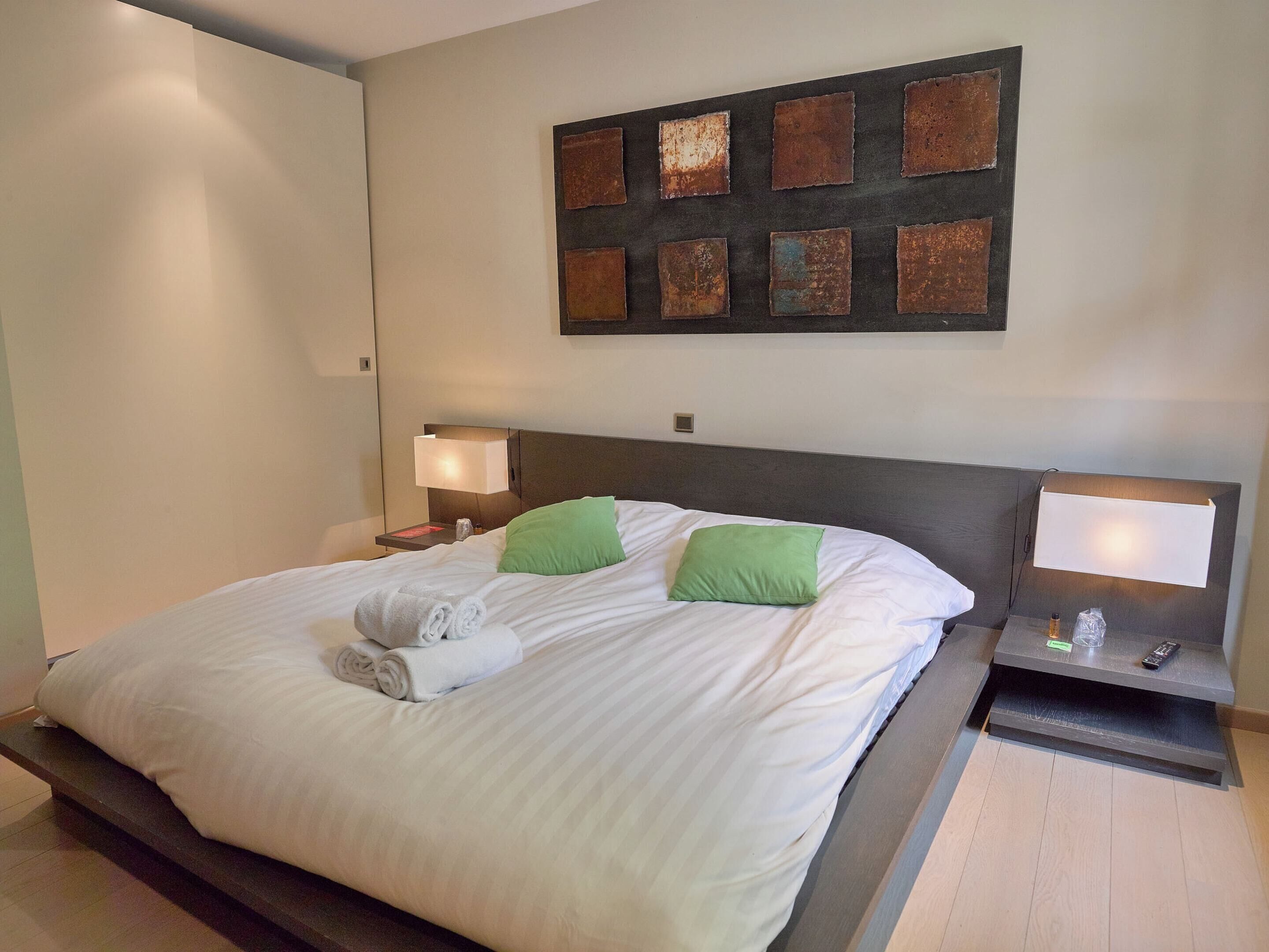 A hotel room with a large bed, two bedside tables, a wall-mounted artwork, and a nightstand with a lamp.
