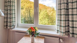 Ein Zimmer mit einem Fenster, das einen bewaldeten Hang zeigt, einem Heizkörper unter dem Fenster und einer Tischvase mit Blumen.