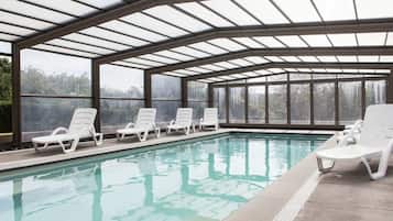 Una piscina techada con paredes de vidrio transparente, rodeada de sillas reclinables blancas.
