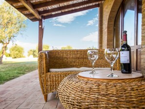 Uma mesa de vime com duas taças de vinho e uma garrafa, sobre um pátio com vista para árvores e um céu limpo.