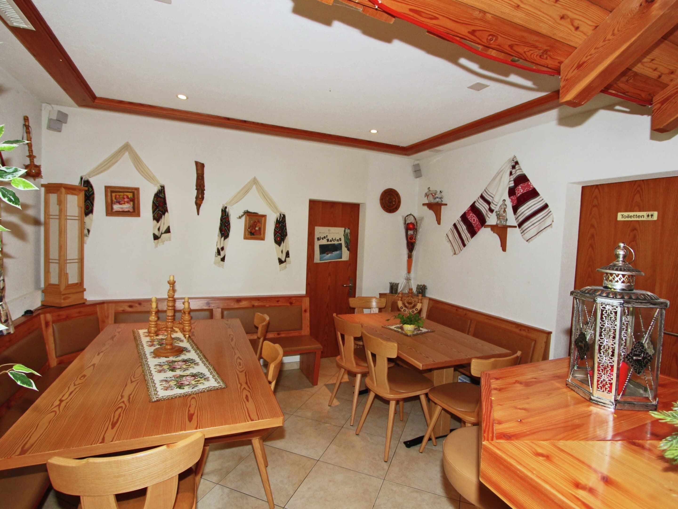 A wooden dining room with a long table and benches, a chandelier, and wall decorations including a lantern and a sign indicating a toilet.