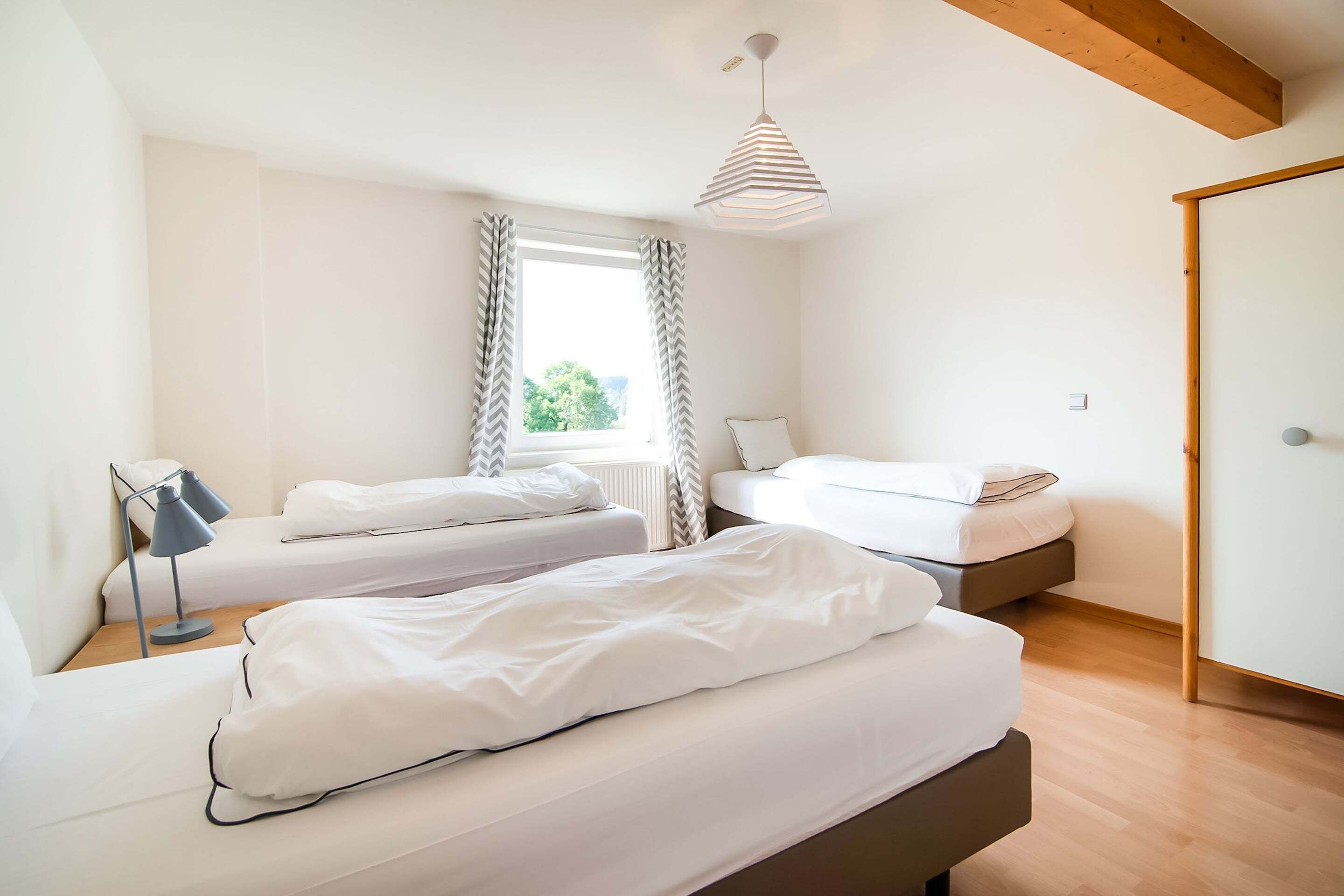Un dormitorio moderno con dos camas individuales, una mesita de noche con lámpara, una ventana con cortinas y una viga de madera en el techo.