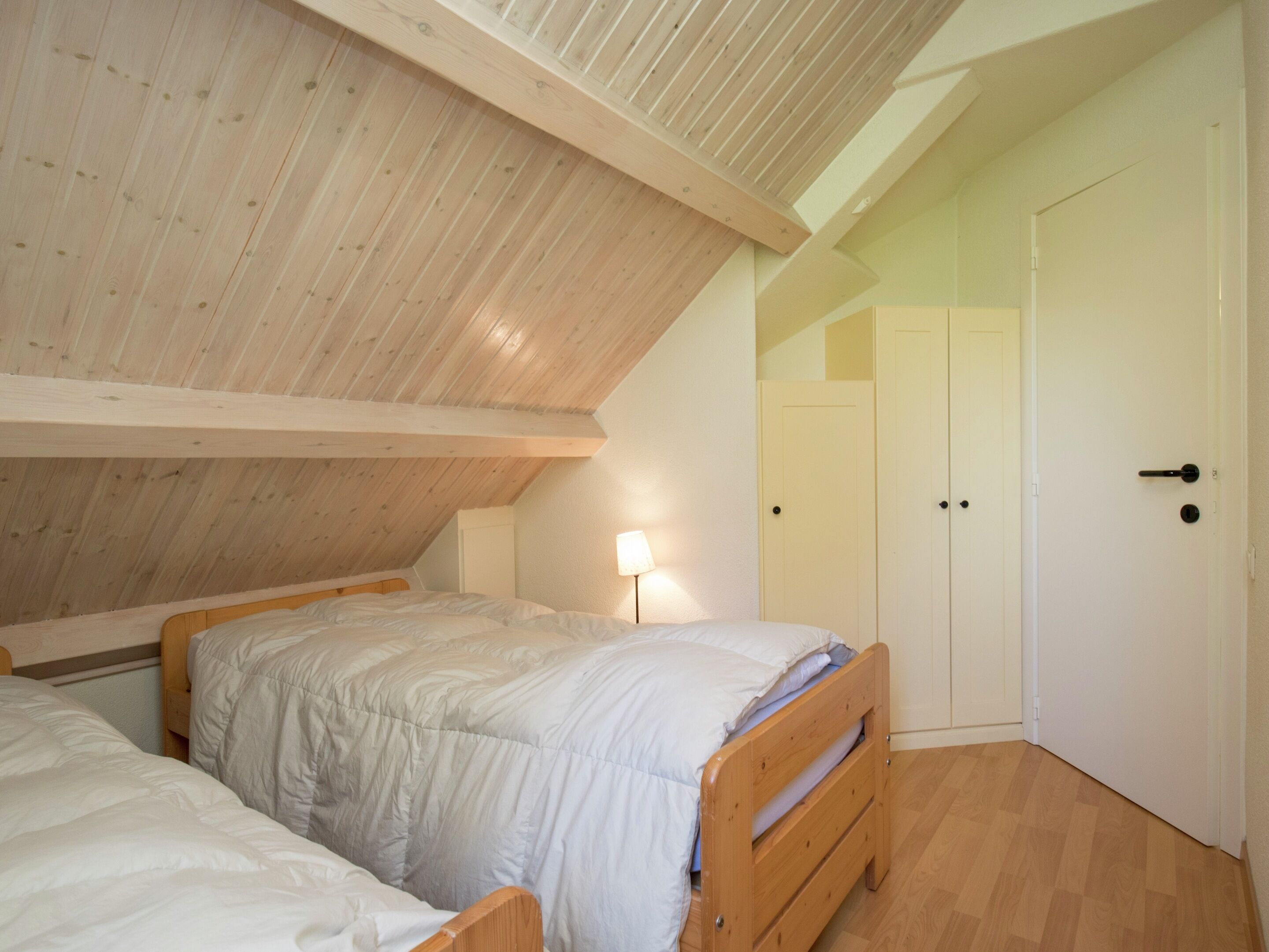 A bedroom with a sloped ceiling, wooden beams, two beds, a nightstand, a lamp, and a wardrobe.