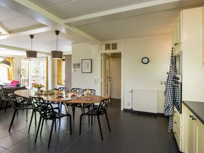 A dining area with a round table, chairs, and a buffet setup. There is a kitchen area with a refrigerator and a clock on the wall. - Rendeux Villa With Garden (Rendeux)