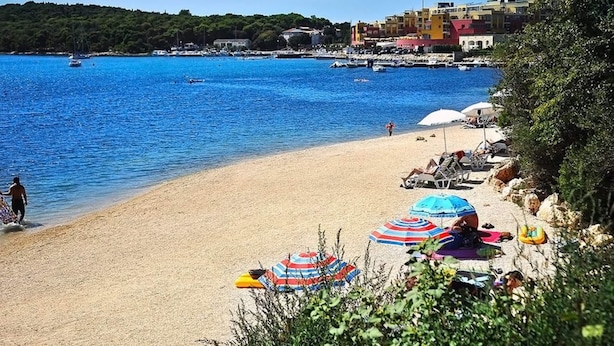 Ein Strand mit klarem, blauem Wasser, Sonnenschirmen und einigen Personen.