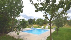 Ein rechteckiger Pool mit Betonumrandung, umgeben von einem Kiesstreifen. Im Hintergrund stehen zwei weiße Liegestühle und eine kleine Gartenhütte.