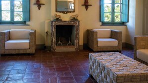 A cozy room with a fireplace, two armchairs, a woven ottoman, and two windows with shutters. - Villa in Tuscany Near Medieval Ghizzano (Peccioli)