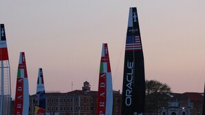 A marina with sailboats, including one with 'ORACLE' and 'PUMA' logos, and another with 'PRADA' and 'Red Bull' logos.