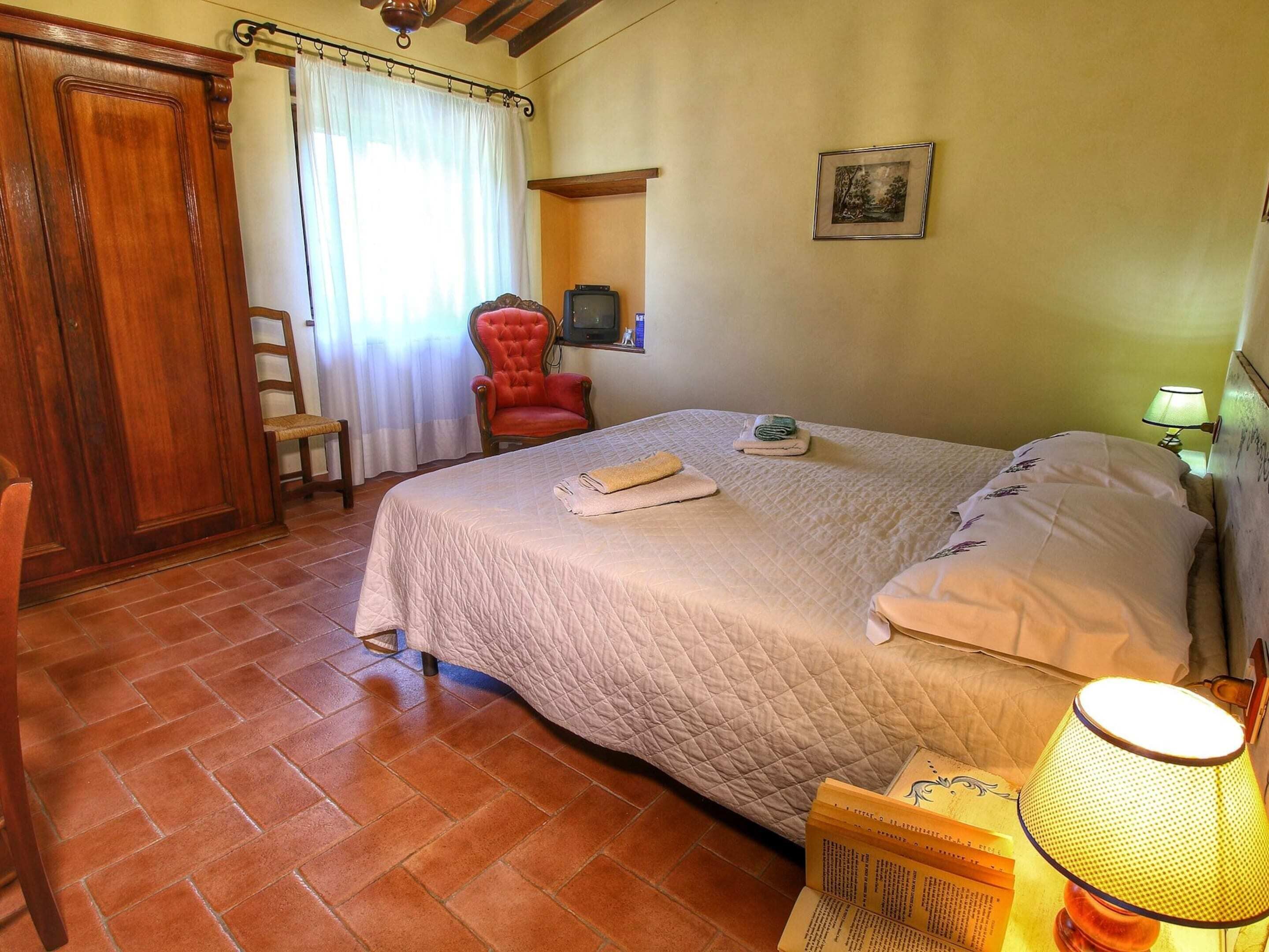 Une chambre à coucher avec un grand lit, une armoire en bois, un fauteuil rouge, une télévision et un tableau encadré au mur.