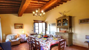 A dining area with a wooden table set for six, surrounded by chairs. A chandelier hangs from the ceiling, and a cabinet with glass doors is to the right. - Escape to Vineyards, Elba (Radda in Chianti)