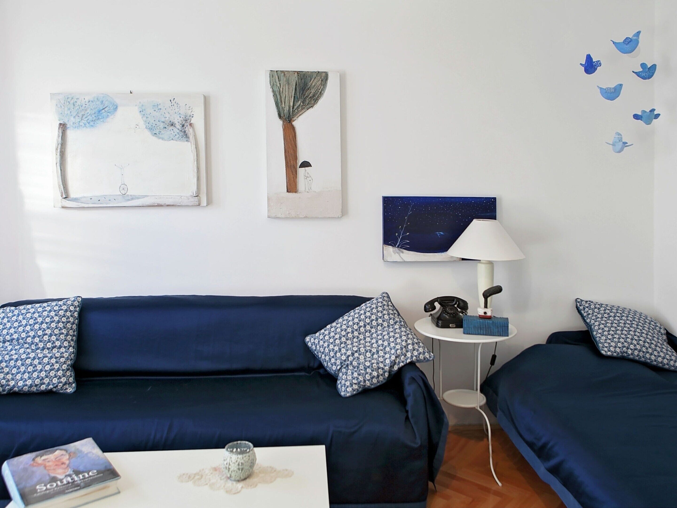 A room with two dark blue sofas, a small round table with a lamp, a book, and wall art including a tree and a night sky.
