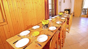 Una larga mesa de comedor de madera dispuesta con platos, vasos y cubiertos, rodeada de sillas de madera en una habitación con paredes de madera y suelo de baldosas.