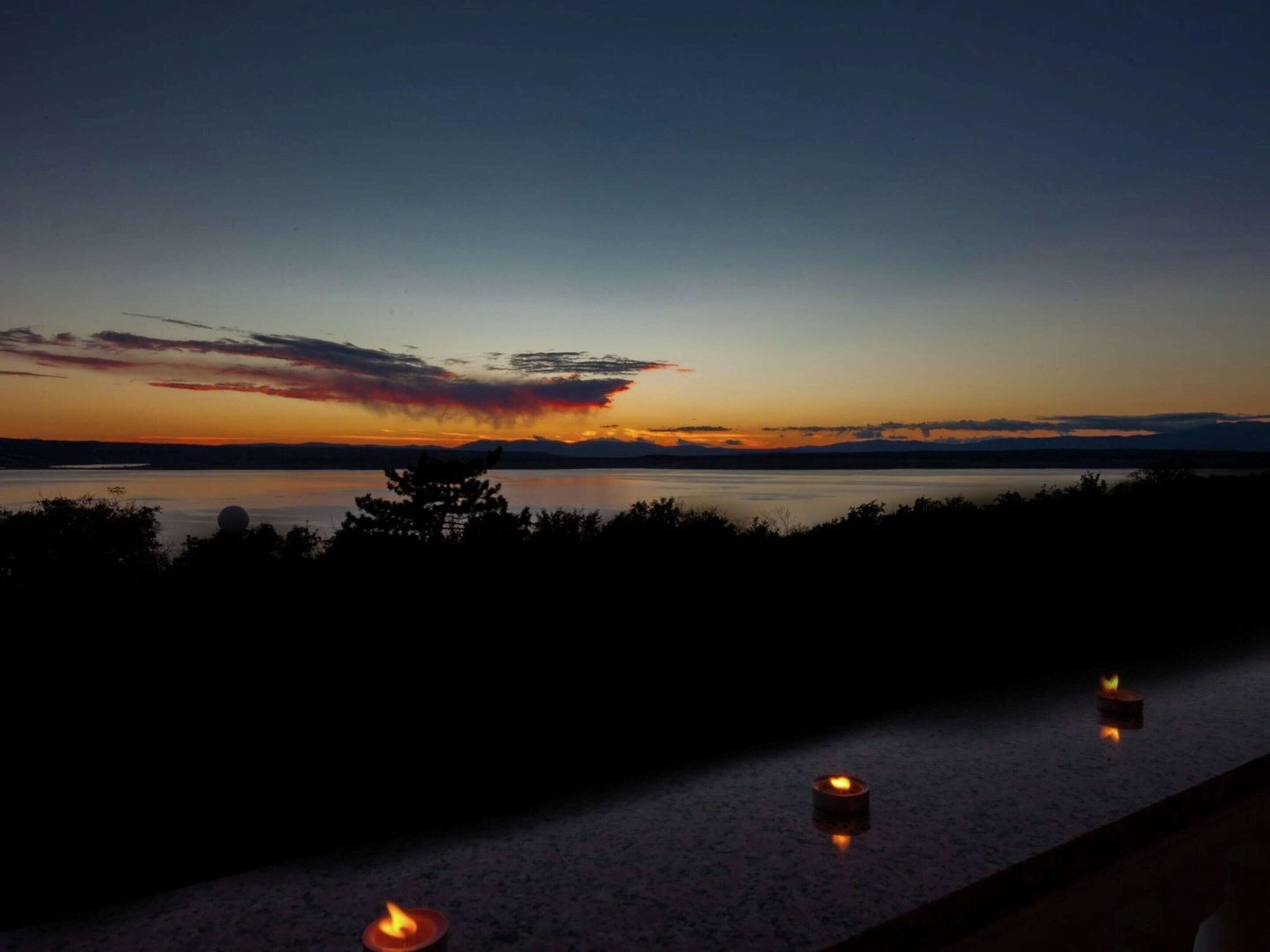 Een zonsondergang vanaf een balkon met uitzicht op een rustig wateroppervlak, met drie brandende kaarsen op de rand.