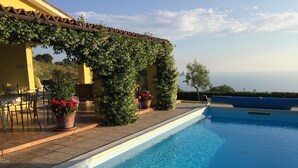 Piscina azul cristalina com um pergolado coberto de vegetação, vasos com flores vermelhas e vista para o céu.