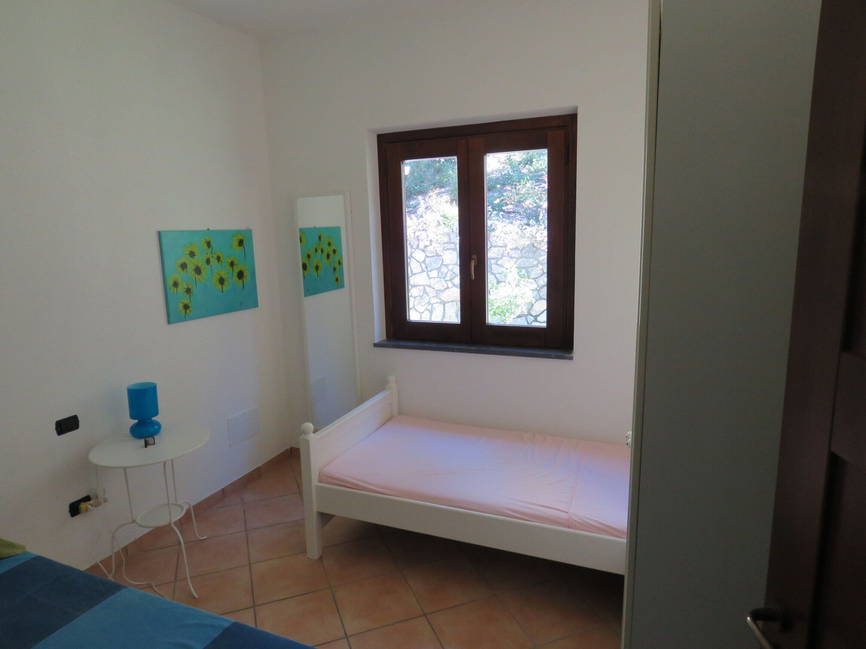 A small, single-bed room with a window, a small table, and two wall-mounted paintings.