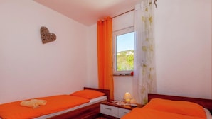 A room with two beds, each with orange bedding, a wooden floor, a small bedside table with a lamp, and a window with orange curtains. - Farm Stay in Croatia With Pool & Forest Views (Crikvenica)