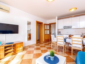 A modern living room with a flat-screen TV, wooden furniture, a dining area with a table and chairs, and a kitchen with white cabinets.