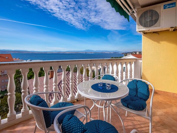 A balcony with a white balustrade, a round table with two chairs, and a view of the sea and distant mountains.