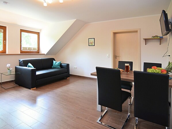 A modern living room with a black sofa, a glass coffee table, and a dining table with chairs. There is a door leading to another room and a TV mounted on the wall. - Holiday Home in Bavaria Near Hiking Trails (Eslarn)