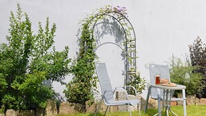 Un jardin avec une chaise et une table en métal, une structure pour plantes grimpantes et un mur en pierre.