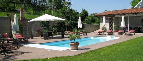 A pool area with lounge chairs and umbrellas, surrounded by greenery and a stone building.