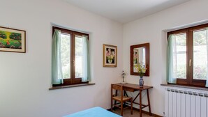 Quarto com cama, escrivaninha, cadeira, janela com cortinas, radiador e um quadro emoldurado na parede.