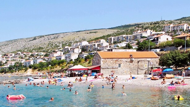 A beach with clear water, people swimming and sunbathing, and buildings in the background. - Apartment in Senj With Sea Views & BBQ (Senj)