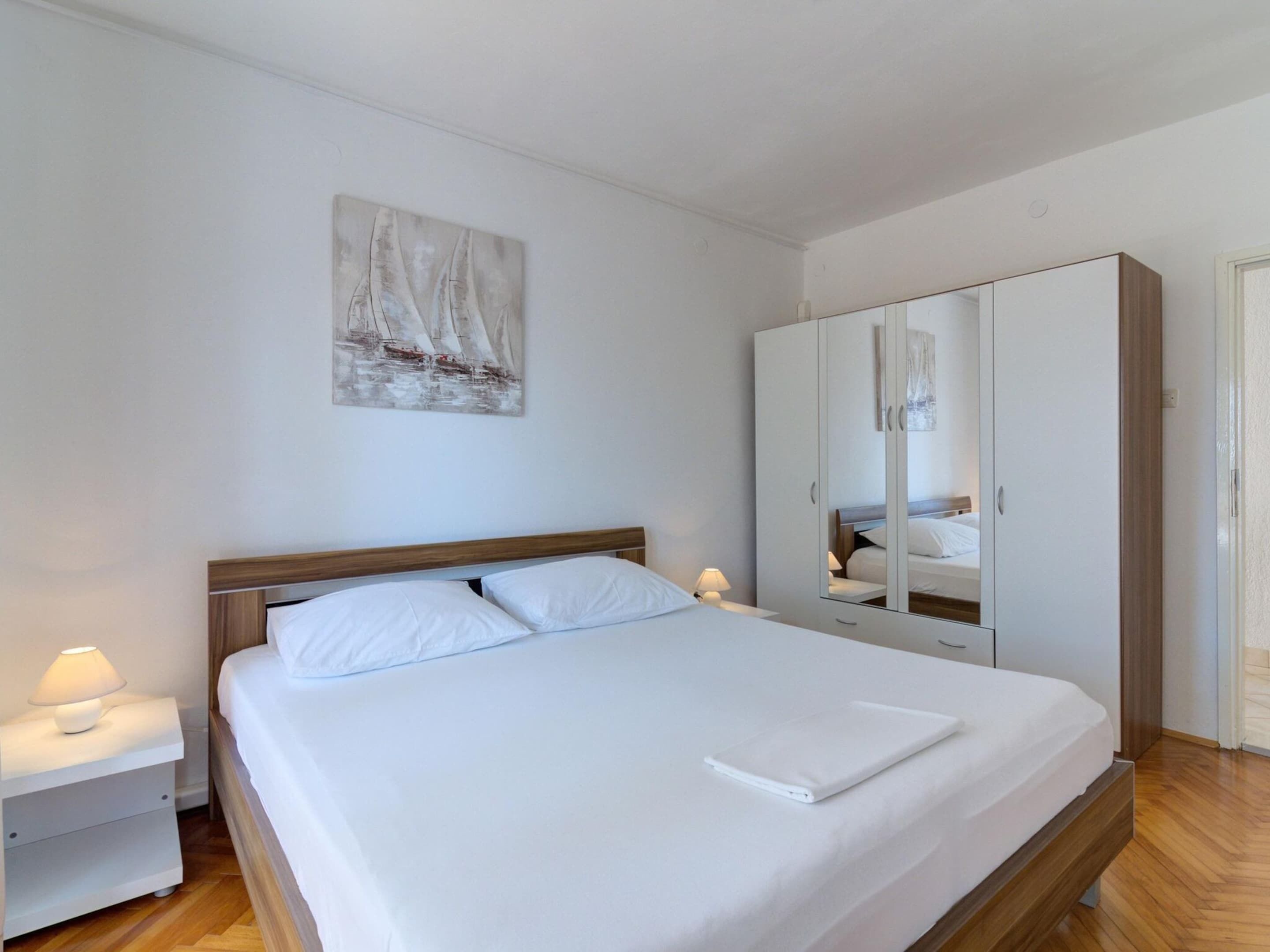A neatly made bed with white linens, a wooden headboard, a bedside table with a lamp, and a large wardrobe with mirrored doors.