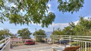 A balcony with two wooden lounge chairs and a table, overlooking a parking area with several cars and a red van. - Apartment in Senj With Sea Views & BBQ (Senj)