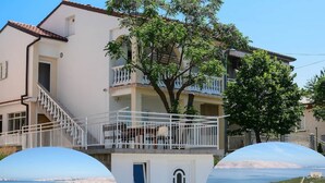 A two-story building with a balcony, a tree in front, and a view of a coastal town. - Apartment in Senj With Sea Views & BBQ (Senj)