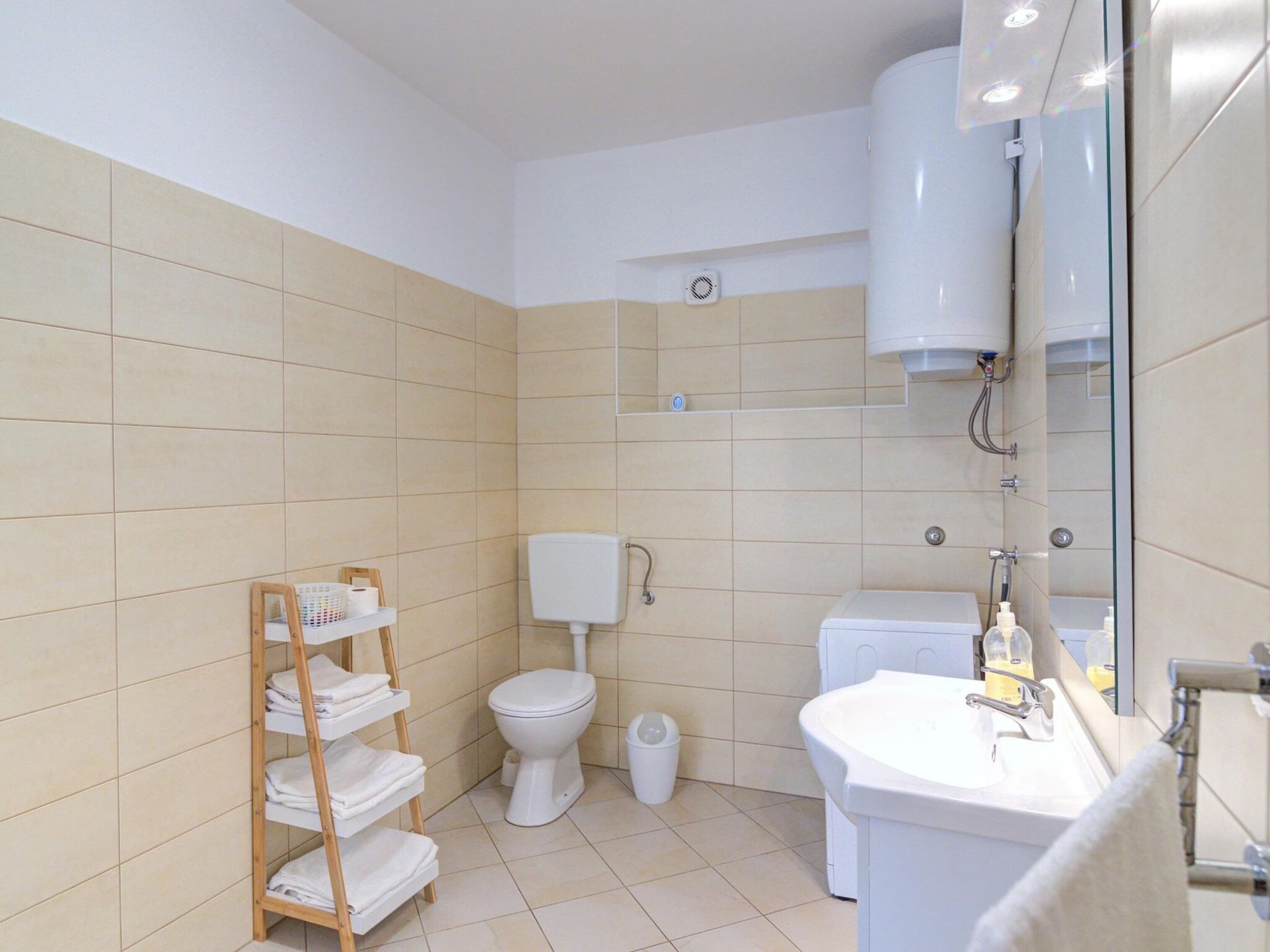 A modern bathroom with a toilet, sink, and shower. There is a shelf with towels and a trash bin.