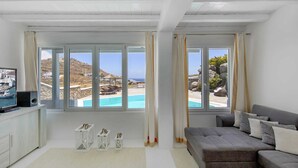 A modern living room with a large window overlooking a pool and a scenic view of a coastal area. - Villa in Mykonos With Sea View and Pool (Mykonos)