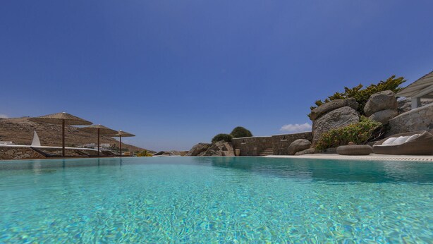 A clear blue pool with lounge chairs and umbrellas, surrounded by rocks and a stone wall. - Villa in Mykonos With Sea View and Pool (Mykonos)