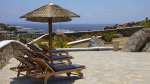 A terrace with wooden lounge chairs and a thatched umbrella, overlooking a coastal landscape with buildings and the sea. - Villa in Mykonos With Sea View and Pool (Mykonos)