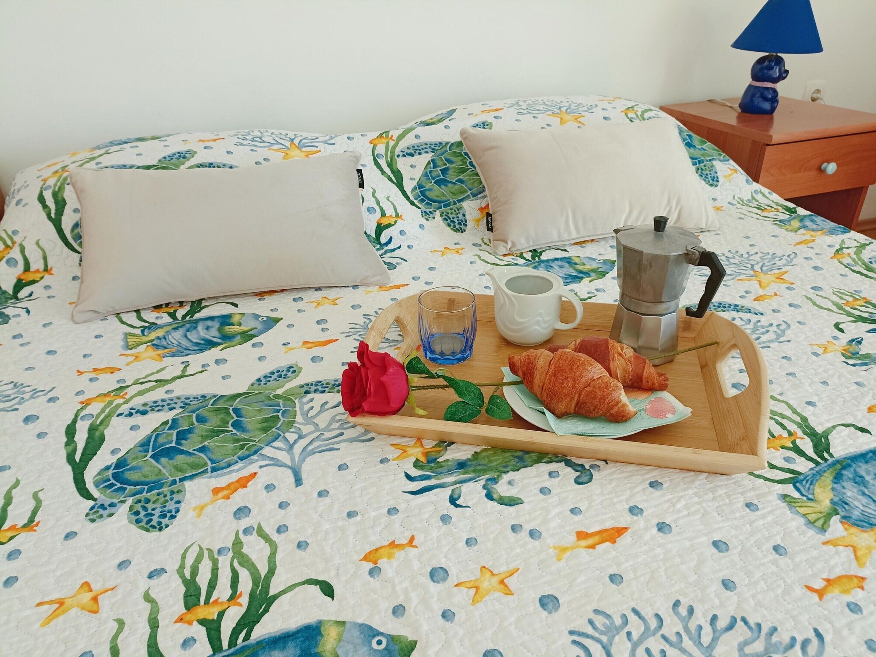 A bed with a colorful aquatic-themed bedspread, a tray with croissants, a coffee pot, a cup, and a glass of water.