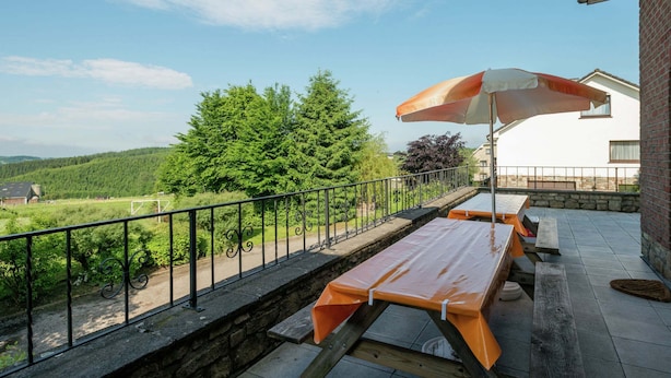 Un patio al aire libre con una mesa y bancos de madera, barandilla de metal y vistas a un paisaje verde con árboles y un edificio al fondo.
