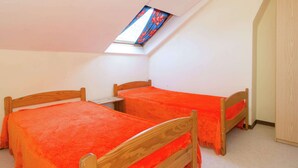 Un pequeño dormitorio en el ático, de forma triangular, con dos camas de madera, un tragaluz y un armario de madera.