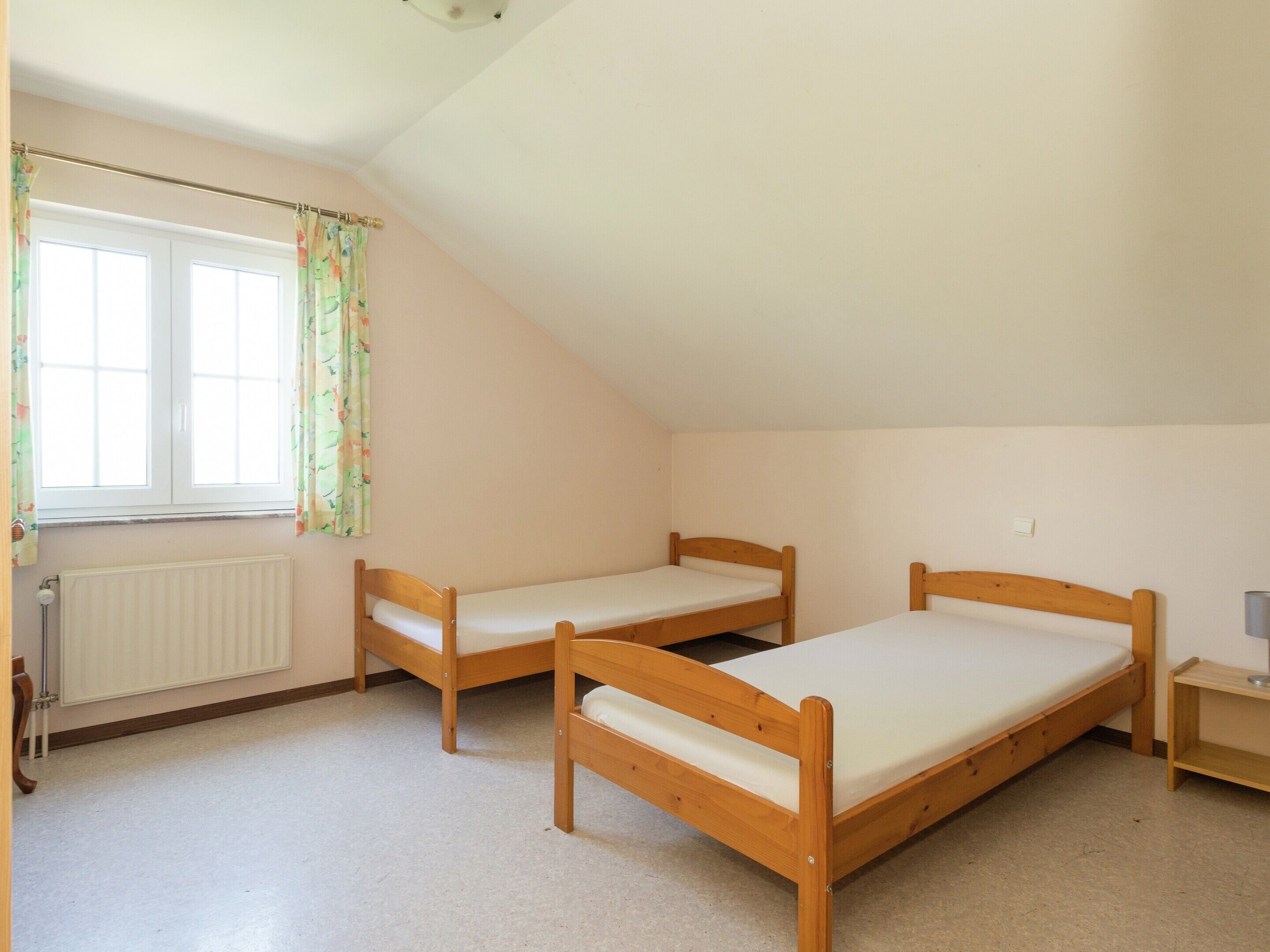 A room with two wooden beds, a radiator, a window with floral curtains, and a small bedside table.