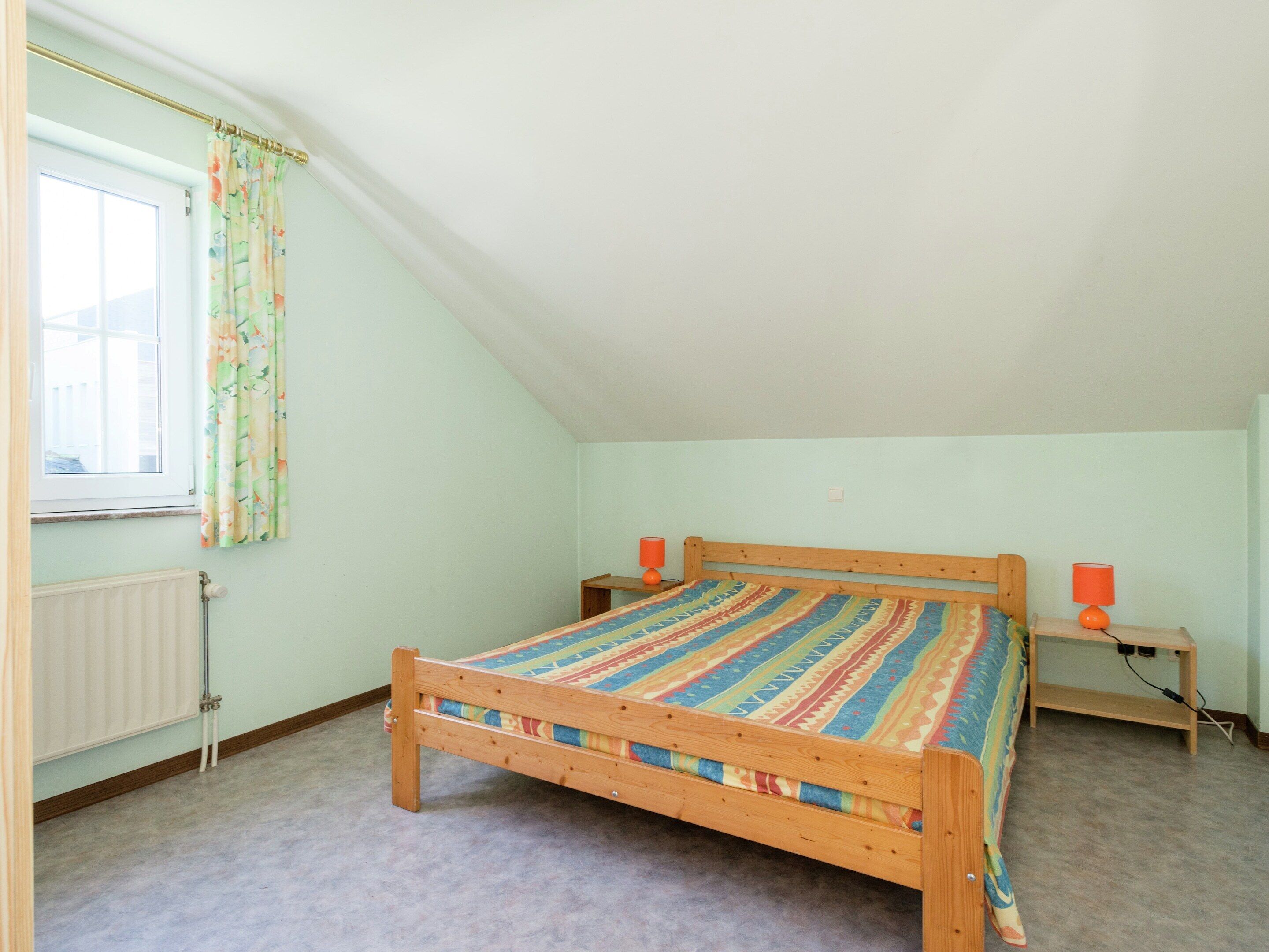 Un dormitorio con techo inclinado, una cama de madera con colcha a rayas de colores, dos mesitas de noche con lámparas, un radiador y una ventana con cortinas estampadas.
