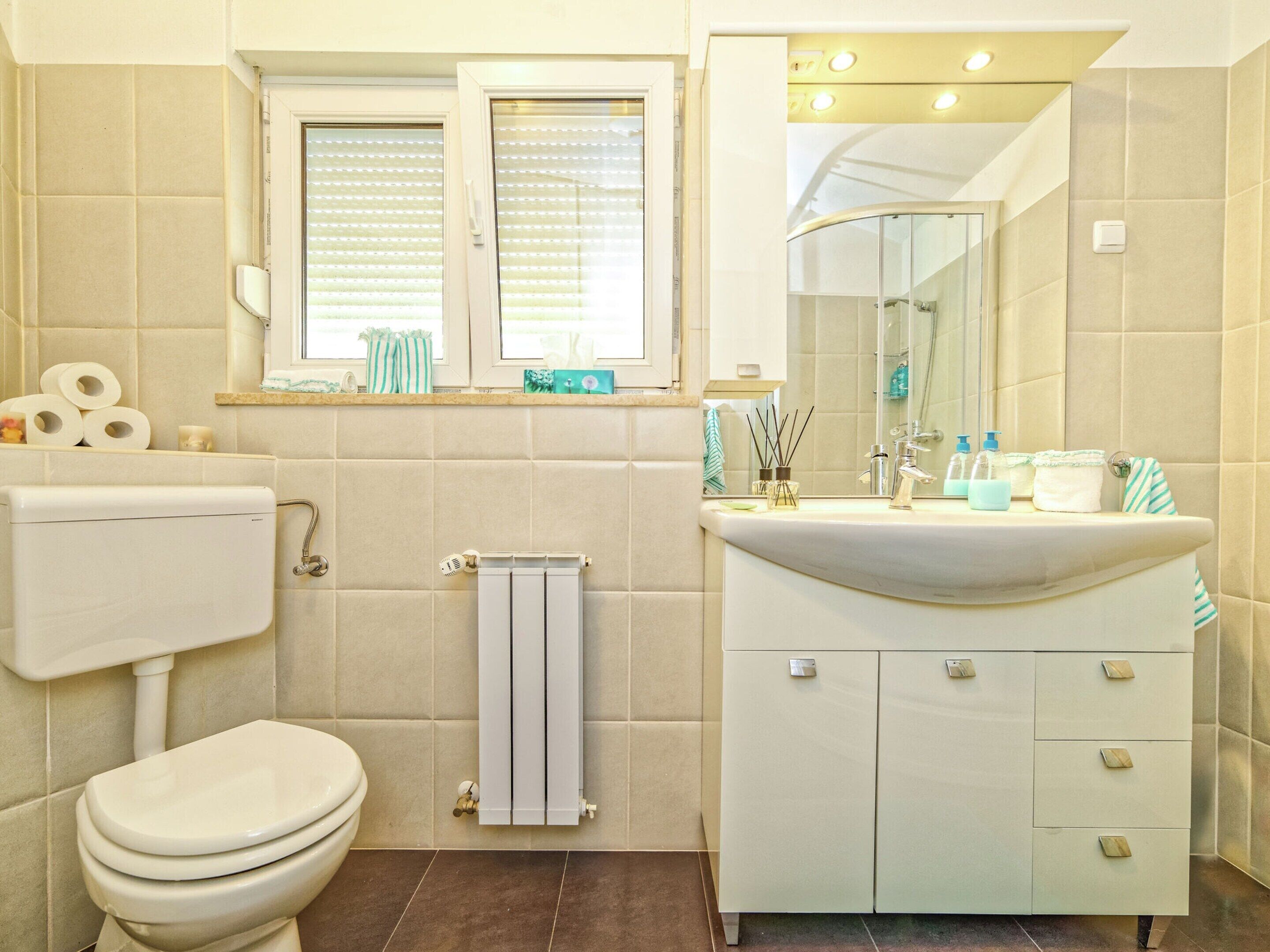 Une salle de bain avec des toilettes, un lavabo avec un miroir et une fenêtre avec des stores.