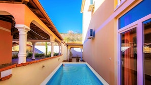Uma piscina estreita com deck revestido, rodeada por um alpendre coberto com colunas e plantas em vasos.