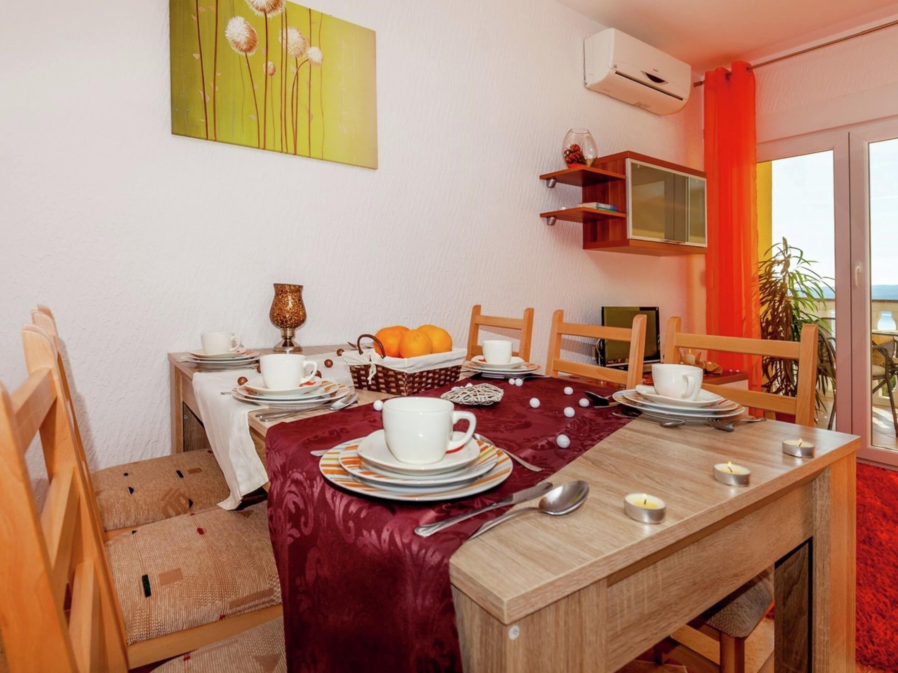 A dining area with a wooden table set for four, surrounded by chairs. There is a red table runner, a white tablecloth, and a painting on the wall. A small shelf with decorative items is above the table.