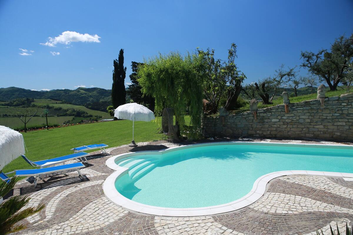 Une piscine bordée de pierres, entourée de chaises longues et de parasols, avec en arrière-plan des collines et des arbres.